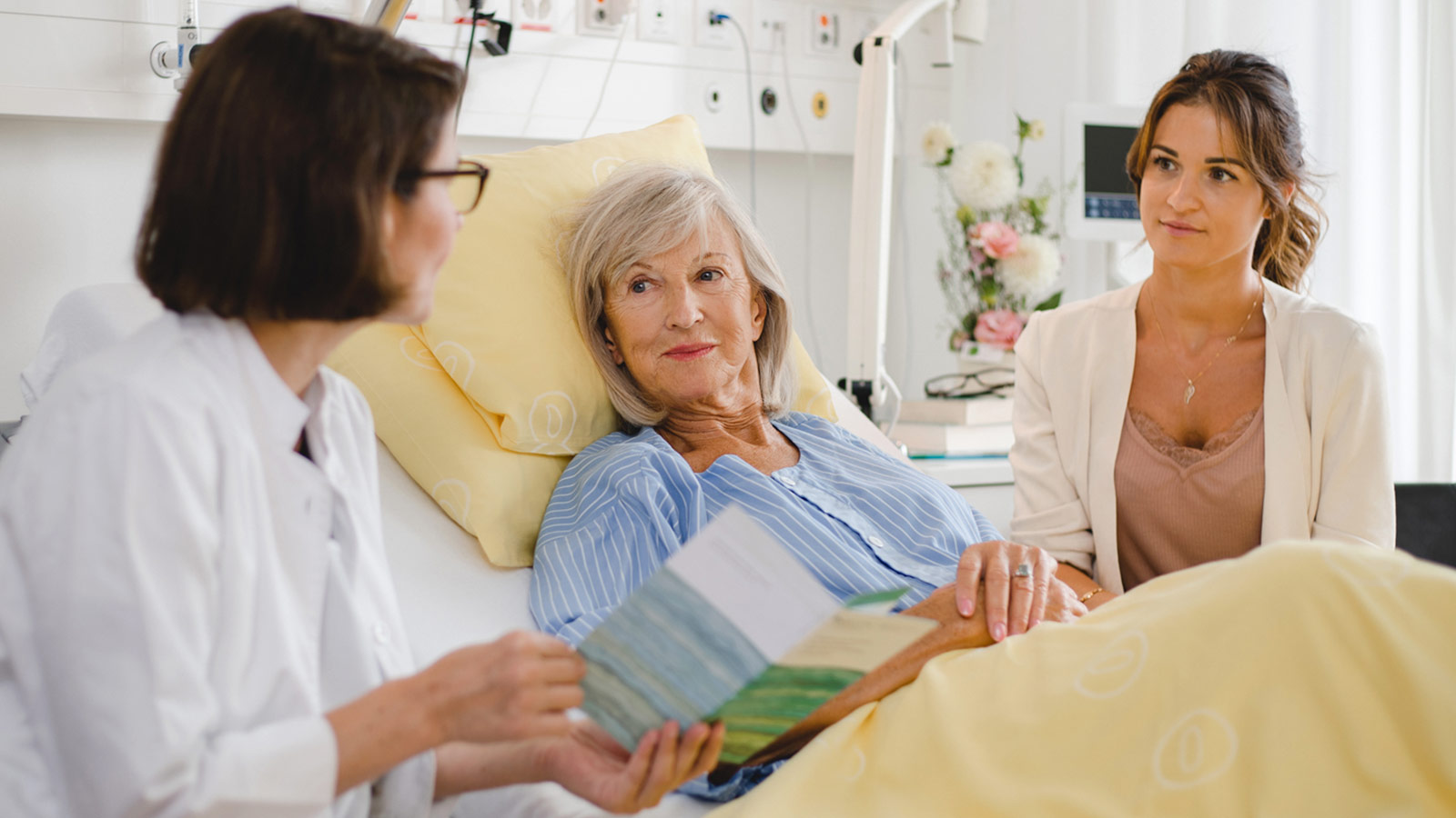 Foto einer im Bett liegenden Patientin mit einer Angehörigen an der Seite, die von einer APN beraten werden.